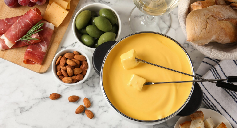 Creamy cheese fondue in a pot with skewers, served with prosciutto, olives, almonds, and crusty bread on a marble table.