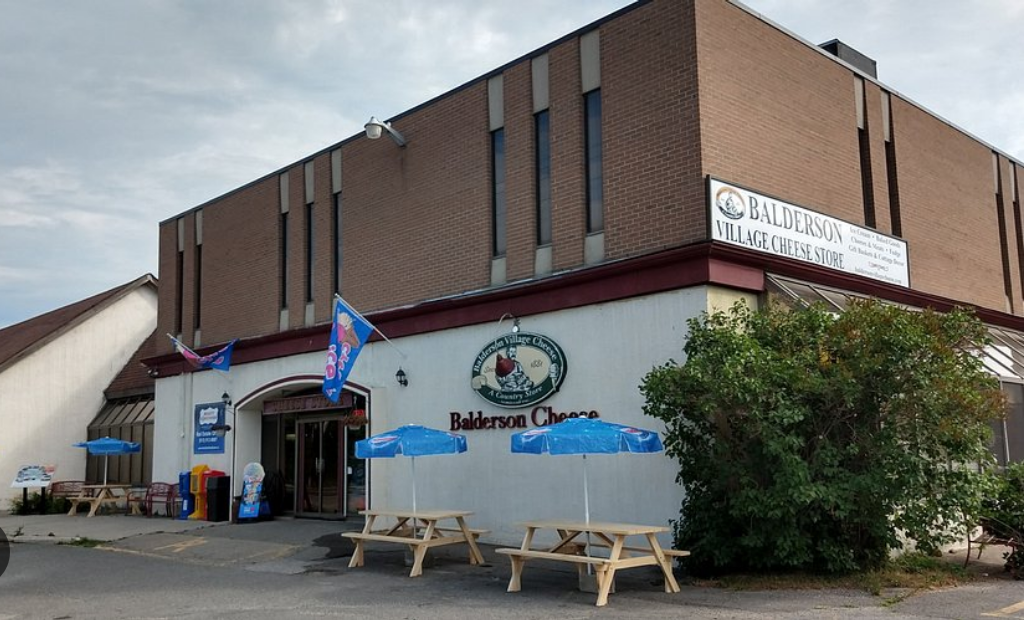 Brick building of Balderson Village Cheese Store with outdoor picnic tables and blue umbrellas on a sunny day.