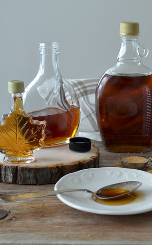 Glass bottles of golden maple syrup on a rustic wood slice with a spoonful of syrup on a white plate.