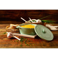 Green ceramic casserole dish with golden baked garlic bread dip, surrounded by bread slices, garlic, and chives.