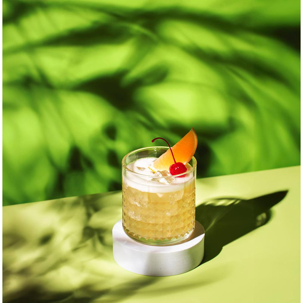Textured glass of golden sour mix cocktail with orange slice and cherry on a white stand.