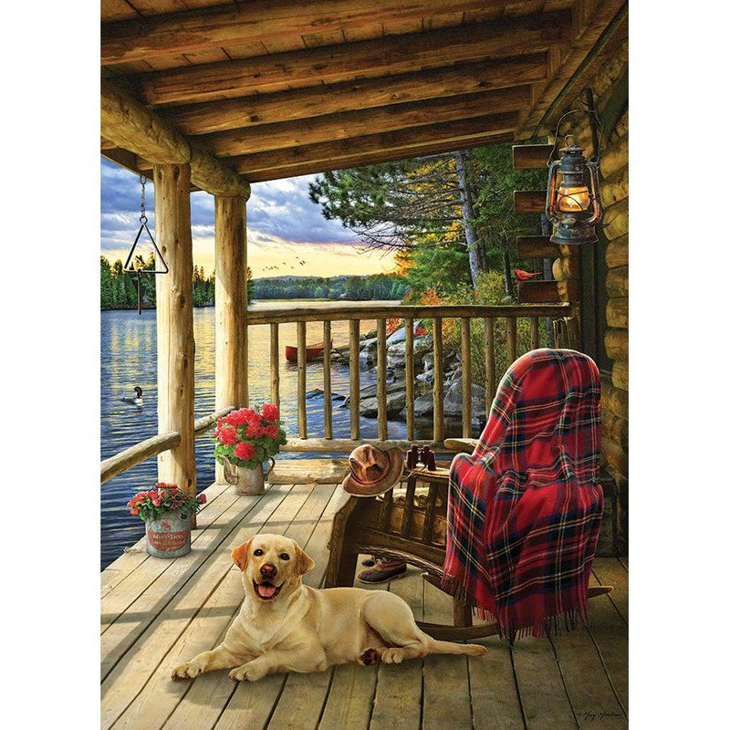 Yellow labrador on a rustic cabin porch overlooking a lake at sunset, with a rocking chair and red plaid blanket.